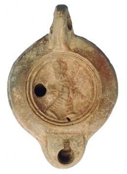 ROMA. Imperio Romano. Lucerna (II-III d.C.). Terracota. Figura masculina coronada avanzando hacia der. En la base un sello y epígrafe. Longitud 10,2 cm. 
